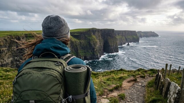  Back view of a female hiker with a backpack admiring the majestic Cliffs of Moher in Ireland. Dramatic coastal landscape with ocean and clouds. AI image