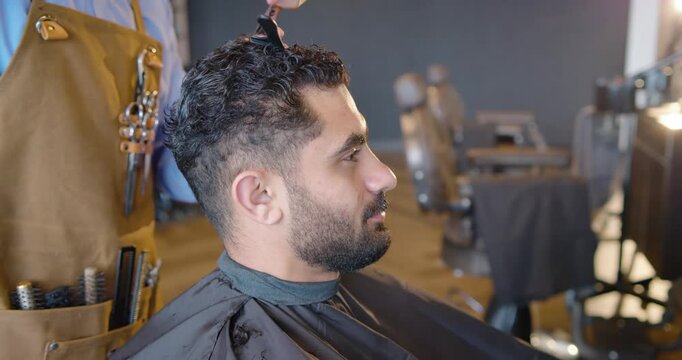 Male barber sectioning male client's hair with comb and clip, scissoring to shorten top at salon