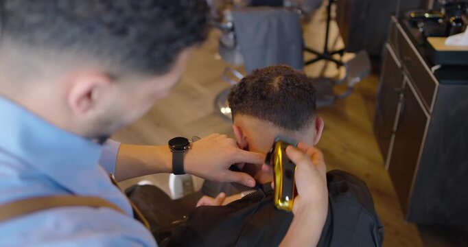 African American barber trimming client hair at shop after head tilt, making fade with gold clipper