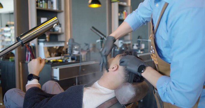 Male client reclining while barber activating steamer, massaging and combing curls for styling