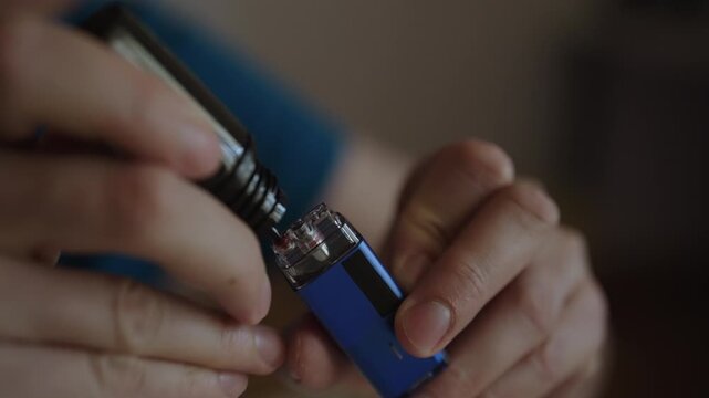 Man hands holding modern electronic cigarette refilling its pod with flavored nicotine e liquid from dropper bottle, preparing device for vaping indoors, close-up. Shooting in slow motion