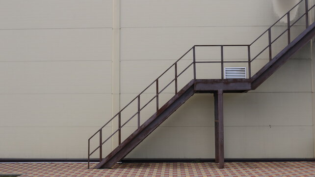 An external metal fire escape is installed along the smooth wall of the building, constructed of beige sandwich panels. The structure consists of inclined flights of stairs, protective railings