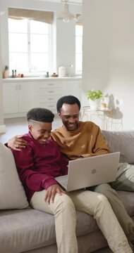 Vertical video: Father and teen son on sofa watching laptop clip, leaning in and laughing