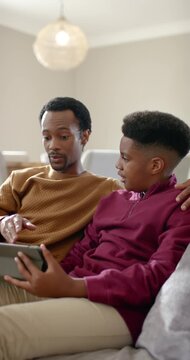 Vertical video: African American father and son leaning on couch, father explaining tablet content
