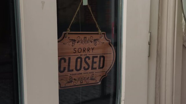 Closed wooden hanging sign displayed behind glass on storefront door, indicating business not open, with rustic decorative lettering, rope hanger, neutral exterior framing and quiet daytime atmosphere