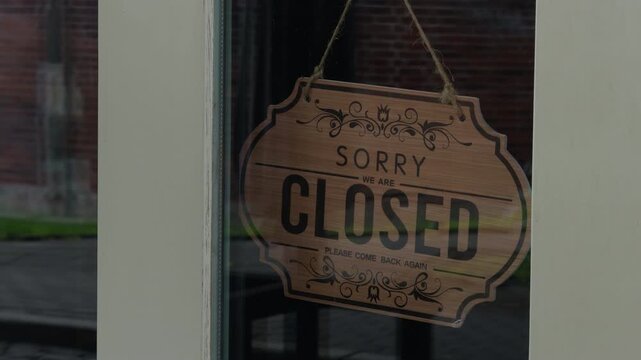 Closed shop sign hanging inside window of small storefront, wooden plaque with decorative lettering on glass, quiet business entrance with muted reflections and empty street outside