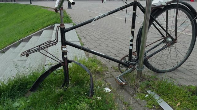 Rusty abandoned bicycle locked to metal post beside paved sidewalk, broken frame, worn wheels, weathered paint, grass, concrete steps, and quiet urban street corner in daylight