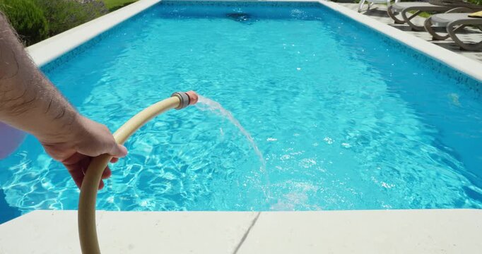Hand holding a garden hose while filling a swimming pool with fresh water. Pool maintenance, water supply, summer preparation, and home outdoor living concept.