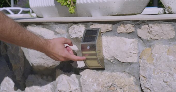 Close-up of a person activating or checking a solar-powered outdoor wall light mounted on a stone wall. Renewable energy, garden lighting, home security, and sustainable outdoor illumination concept.