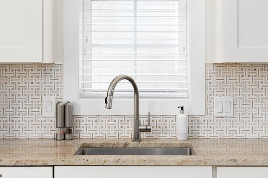 A kitchen faucet detail with a stainless steel faucet and sink, brown and white basketweave tile, brown granite countertop, and cream cabinets.