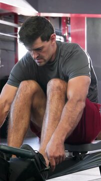 Vertical video: Entering frame, man in red shorts strapping feet on erg in gym, rowing for cardio