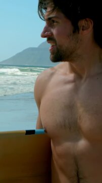 Vertical video: Seeing waves, shirtless surfer holding yellow board in red trunks preparing to surf
