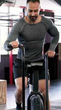 Vertical video: Starting pedaling, mid-adult man powering fan bike in gym in grey shirt for cardio