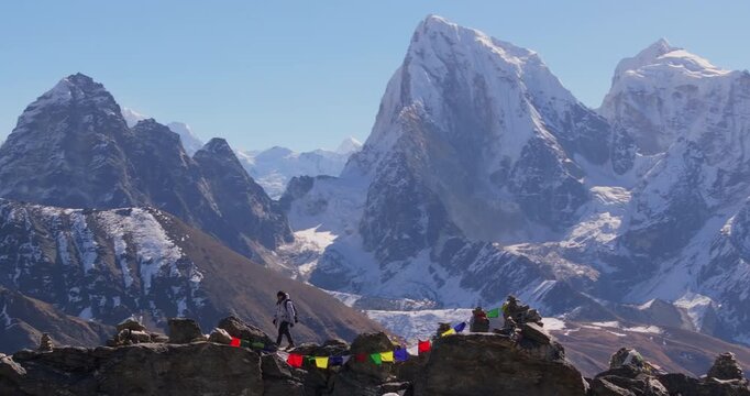 Gokyo Ri, Everest Region Nepal, drone orbits a man walking past a small stone stupa and colorful prayer flags along a rocky mountain ridge with the Himalayan mountain range in the background.