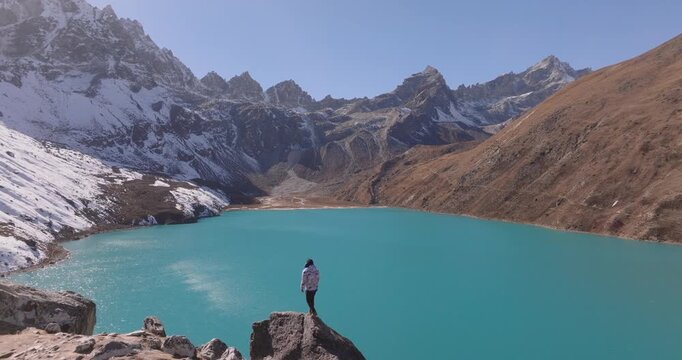 Gokyo region, Nepal, aerial view of a lone hiker standing on a rock outcrop overlooking a vast turquoise alpine lake surrounded by snow-capped peaks and barren slopes under a clear blue sky.