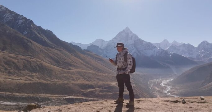 Thugla Pass Everest Base Camp trekking route Nepal, aerial drone orbits a male trekker speaking to a walkie talkie radio in a wide valley, winding river, and the towering mountain peaks of Ama Dablam