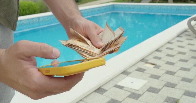 A man at a poolside during vacation holding cash and a smartphone, representing unexpected roaming charges and mobile costs while traveling abroad.