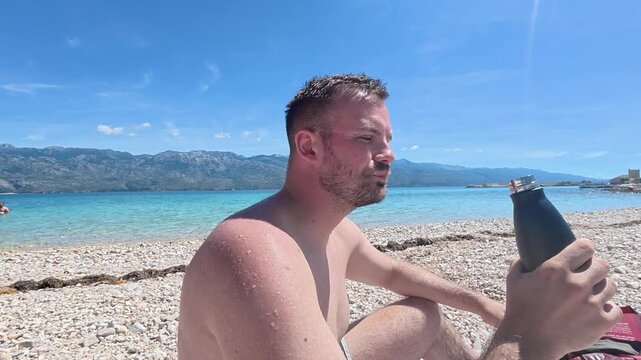 Man drinking from a water bottle while relaxing at the beach on a hot summer day. Hydration, healthy lifestyle, heat protection, and dehydration prevention concept.