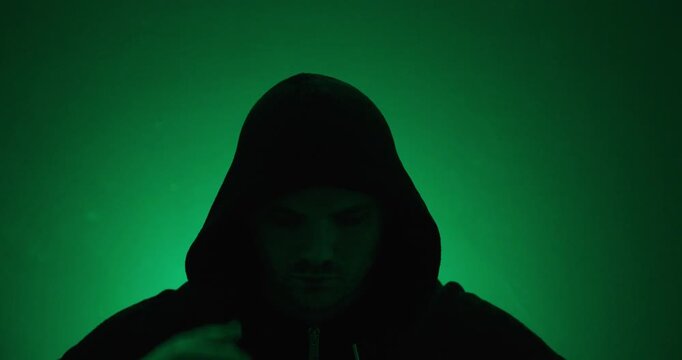 Slow-motion shot of a man pulling up the hood of a hoodie under green dramatic lighting. Mystery, anonymity, suspense, cinematic atmosphere, and dramatic portrait concept.