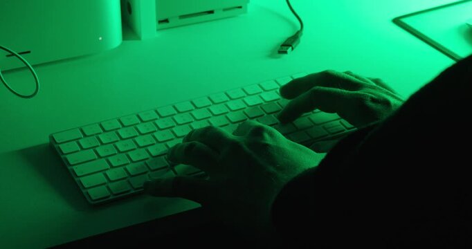 Close-up of hands entering the frame and typing on a computer keyboard under dramatic green lighting. Technology, programming, office work, productivity, and digital workspace concept.