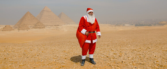 Fototapeta premium Whimsical Santa Claus in red costume stands holding a gift sack before the Giza pyramids in the sunny Egyptian desert ideal for cross cultural holiday