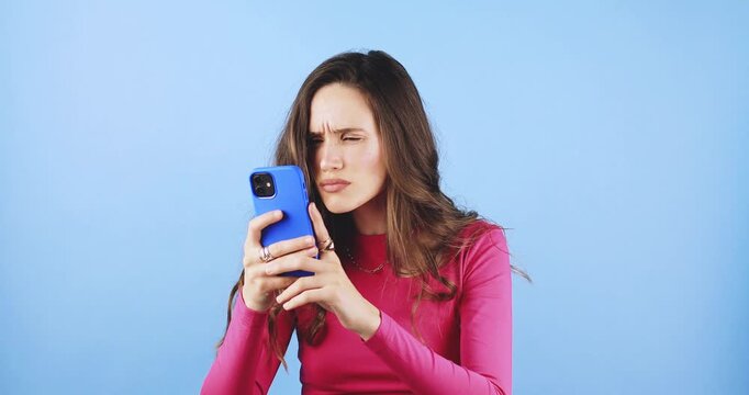 Young woman looking at smartphone screen with confused, skeptical, and annoyed facial expression, browsing content with frown studio isolated on blue background, concept of negative digital experience