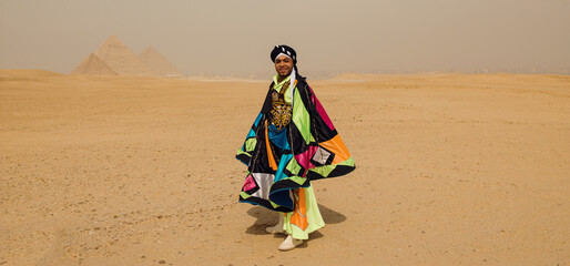 Fototapeta premium Joyful male Egyptian folk dancer in a bright multicolored tanoura costume with flowing cape standing in the desert before the Giza pyramids ideal for