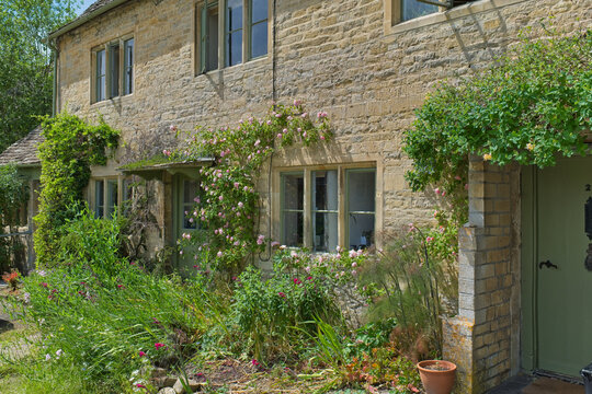 A picturesque stone house with green climbing plants and a vibrant front garden under a sunny sky. The traditional stone walls feature multiple windows in Bibury - Gloucestershire - UK