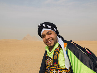 Fototapeta premium Cheerful Egyptian folk dancer in colorful embroidered tanoura costume and black turban smiling warmly in the desert near the Giza pyramids ideal for
