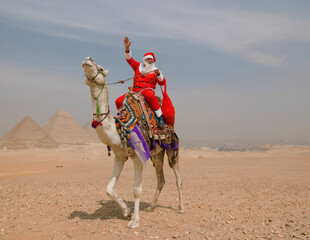 Fototapeta premium Festive Santa Claus waving cheerfully while riding a colorfully decorated camel before the Giza pyramids in the Egyptian desert ideal for holiday