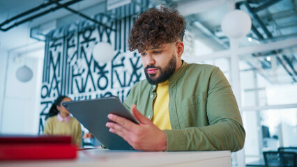 Fototapeta premium Open office interior with abstract wall art. Handsome Caucasian man resting chin on hand. Looking to side with thoughtful expression. Sitting at desk. Considering reply and planning response.