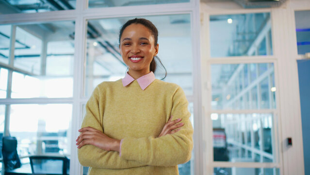 Smiling business woman standing with folded arms near glass wall. Looking at camera with confidence. Modern corporate interior around. Professional female presenting leadership and career success.