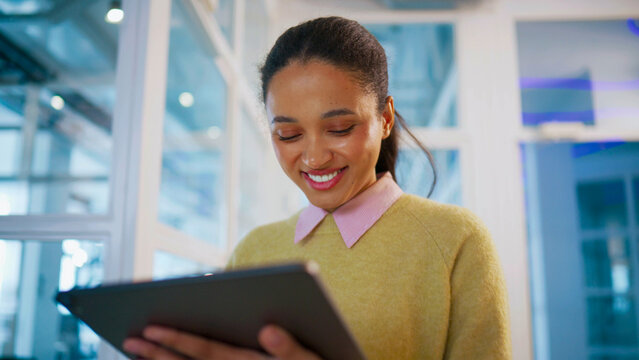 Business woman holding tablet and smiling at screen. Walking along bright office corridor. Female receiving positive message from colleague. Corporate communication and success concept.