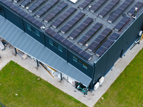 Aerial view of a modern industrial building with a large array of solar panels on its flat roof, situated on a green lawn in Ikskile, Ogre Municipality, Latvia.