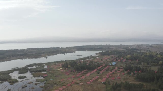 A stunning aerial view of Lake Sevan in Armenia, featuring a winding sandy peninsula, small lakeside settlements, and marshy wetlands under an overcast, cloudy, and atmospheric autumn sky.

