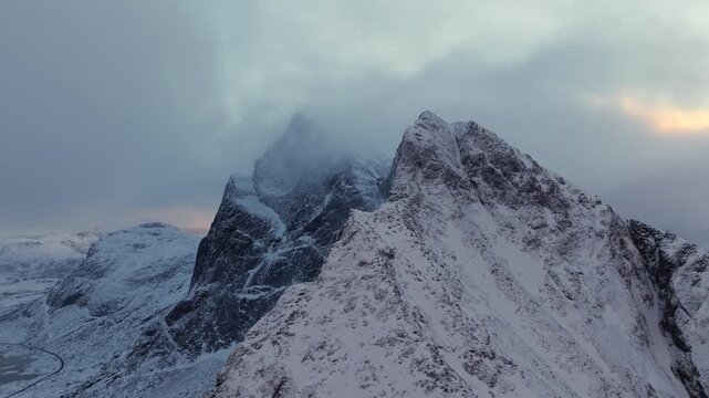 Aerial view of snow-covered jagged mountain ridge with misty peak and winding valley road