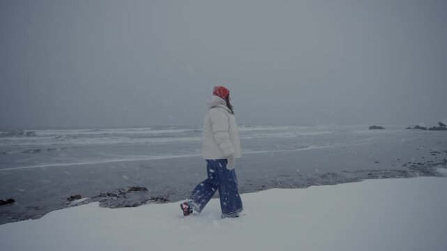 Solitary figure in red hat on snowy shoreline facing stormy grey sea