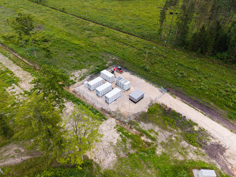 Aerial view of a battery energy storage system with white container units on a gravel pad surrounded by forest in Suntazi, Ogre Municipality, Latvia.