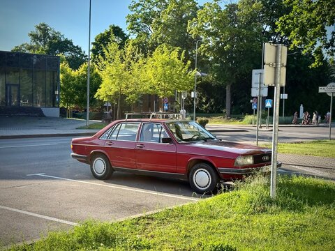 Red Audi 100 C2 sedan parked in an urban street setting. The car features a boxy design typical of the late 1970s and early 1980s, with four doors and a roof rack. 