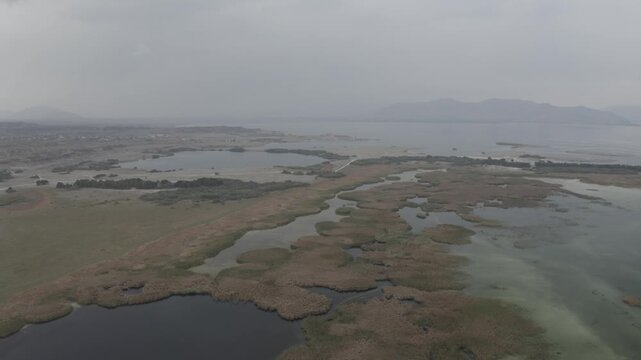 A stunning aerial view of Lake Sevan in Armenia, featuring a winding sandy peninsula, small lakeside settlements, and marshy wetlands under an overcast, cloudy, and atmospheric autumn sky.

