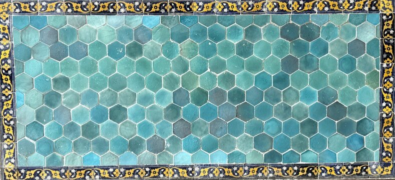 Close-up of a traditional Persian ceramic tile panel serving as a textured background, featuring a honeycomb pattern of turquoise hexagonal tiles framed by an ornate yellow and black floral border.