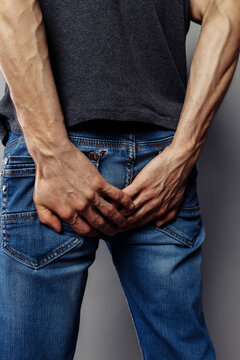 A young, thin guy in jeans is squeezing his ass tightly with his hands. Concept: Queue to the bathroom, intestinal and stomach diseases. expressive veins on the arms