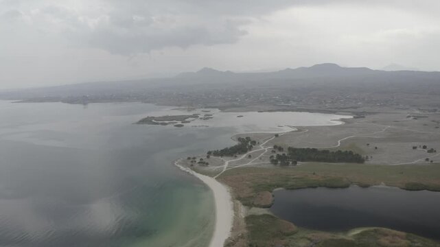 A stunning aerial view of Lake Sevan in Armenia, featuring a winding sandy peninsula, small lakeside settlements, and marshy wetlands under an overcast, cloudy, and atmospheric autumn sky.

