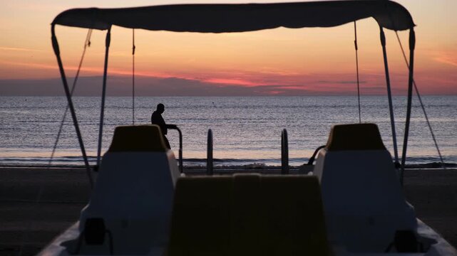 Peaceful 4K video of a silhouetted man walking along the seashore at sunrise with a pedal boat in the foreground, calm water, gentle waves and colorful morning sky.