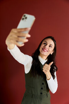 Woman taking real time selfie using smartphone in studio, showing thumbs up gesture, smiling, sharing good feedback experience, red background