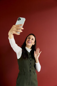 Happy woman recording engaging social media content or participating in a cheerful video call, holding a mobile phone and waving hand on a red background