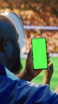 Portrait video: Close-up of mockup smartphone used by man in soccer stadium stands to compare match odds, with football supporter checking live score application predictions on chroma key phone.