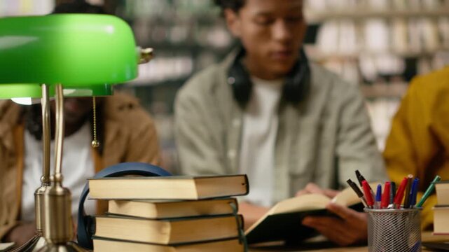 Students working around laptop and open books during library study session, diverse classmates sharing ideas near books, notebooks and green lamps on wooden table, for education technology.