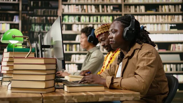 Smiling black student wearing headphones and typing at desktop monitor with books and classmates nearby in warm library studying space for coursework, digital learning, supporting coursework.