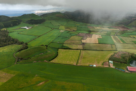 Aerial view of lush green fields and rolling hills of Sao Miguel Island, with low-hanging clouds over the volcanic landscape and distant coastline in the Azores, Portugal.